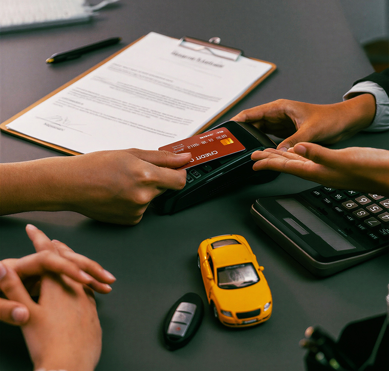 Hands processing a credit card payment at a desk with a yellow toy car and documents, representing a vehicle purchase or service booking.