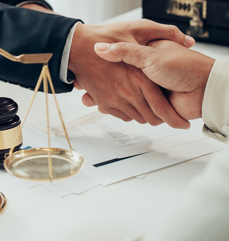 A close-up of a firm handshake between two individuals over legal documents, with a golden scale of justice and a judge's gavel visible in the foreground, symbolizing a successful legal settlement or partnership agreement.